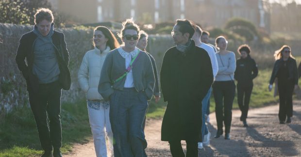 A group of people walk along a road on a sunny day. It's a large group so the number stretch back and people are talking to each other in small groups.