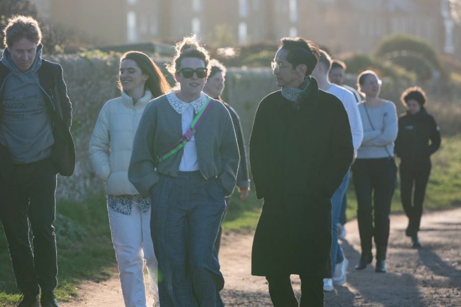 A group of people walk along a road on a sunny day. It's a large group so the number stretch back and people are talking to each other in small groups.