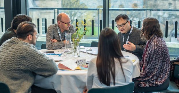 A group of people sit round a table in a well lit room. They're listening to one member of the group who is talking.